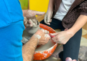 Uczennica usiadła na kole gancarskim naprzeciwko pana i razem z nim lepi swoje naczynie, które kształtem przypomina nie duży wazonik.