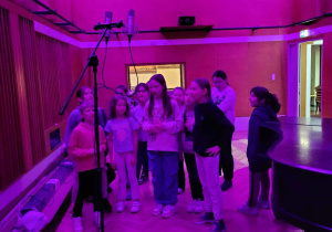 Studio nagrań dźwiękowych - dzieci nagrywają utwory wokalne za pomocą stereofonicznej pary mikrofonów Neumann U87 na uchwycie umieszczonym powyżej ich głów. W tle ustawione światło różowe dla wzmocnienia efektu. Za dziećmi jest małe prostokątne okienko do reżyserni dźwiękowej, czyli pokoju kontrolnego – pomieszczenia z mikserem audio i komputerem do rejestracji i montażu nagrań wykonywanych w studio.