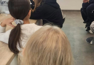 Na zdjęciu widać salę, w której odbywają się warsztaty. Uczniowie siedzą przodem do ekranu i uważnie oglądają wyświetlany slajd. Na ekranie wyświetla się slajd "Zadanie 1. Co mieszka w leśnej ściółce? Przyjrzyjmy się bliżej". Obok tekstu znajduje sie zdjęcie Ziemi.