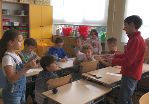 Adam, stojąc przy ławkach uczniów z klasy 2a, tłumaczy i pokazuje, jak składać papier.