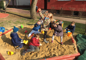 Tymek, Hubert, Kacper, Tonia i Ela wygłupiają się w piaskownicy. Natomiast za nimi chłopcy tworzą budowle z piasku.