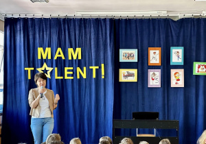 Pani Kierownik zapowiada początek wydarzenia. Na niebieskiej kotarze wisi napis "Mam Talent" oraz kolorowe obrazki.