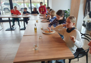 Uczniowie siedza przy dużym stole i jedzą przygotowane przez siebie pizze.