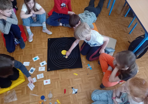 Dzieci siedzą na podłodze, przed nimi leżą elementy do wykonania pracy. Jeden z uczniów przykleja planety do planszy.
