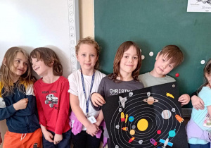 Dzieci stoją przy tablicy i prezentują gotową pracę "Układ Słoneczny" na której znajdują się planety i statki kosmiczne.