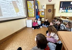 Uczniowie siedzą przed tablicą, oglądają wyświetloną prezentację i czytają podane informacje.