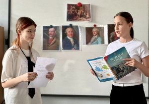 Dwie uczennice stoją przy tablicy, na której wisza zdjęcia Króla Stanisława Augusta Poniatowskiego i prezentują swój pokaz.
