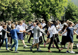 Uczniowie i nauczyciele, ubrani w białe bluzki, tańczą Poloneza na szkolnym boisku.