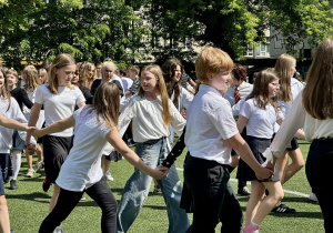 Uczniowie, ubrani w białe bluzki, tańczą Poloneza na szkolnym boisku.