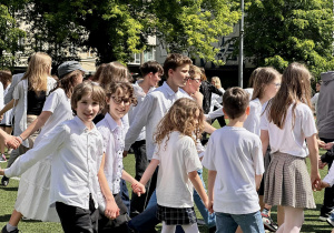 Uczniowie, ubrani w białe bluzki, tańczą Poloneza na szkolnym boisku.