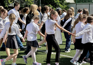 Uczniowie i nauczyciele, ubrani w białe bluzki, tańczą Poloneza na szkolnym boisku.