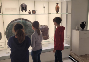 Uczniowie stoją przed szklaną gablotą i patrzą na starożytną tarczę, która chroniła wojownika.