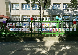 Napis "Trzymamy kciuki za naszych ósmoklasistów" ułożony z kolorowych liter i ozdobiony balonami wisi na ogrodzeniu szkoły.