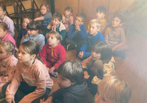 Uczniowie siedza ną podłodze i słuchają opowieści w starej pracowni stolarskiej jak kiedyś wyglądała praca stolarza oraz jakich narzędzi używał.