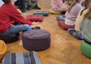 Dzieci siedzą na siedziskach na podłodze i słuchają pani pracującej w bibliotece.