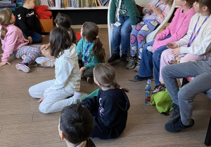 Uczniowie siedzą na podłodze, na krzesełkach w bibliotece i patrzą na zbiór książek na regale.