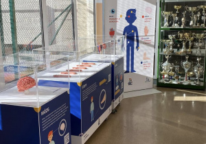 Plakat informujący o tematyce wystawy i gabloty stoją na łączniku prowadzącym do sali gimnastycznej.