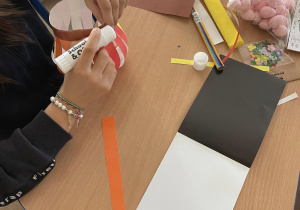 Uczennica wykonuje stroik wielkanocny. Na ławce widać kolorowy papier techniczny, różowe jajeczka styropianowe, różowe piórka, nożyczki i klej.