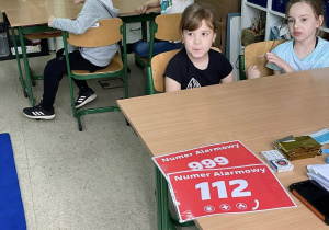 Dzieci siedzą przy stołach i biorą udział w szkoleniu z udzielania pierwszej pomocy. Na początek poznają teorię.