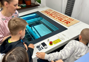 Uczniowie stoja przy ploterze laserowym. Chłopiec, na prośbę Pani, włącza filtr, który włącza się przed rozpoczęciem obróbki laserowej.