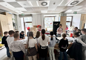 Uczniowie znajdują się w stolarni i stoją przy stole do stolarskim.