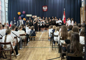 Pani Dyrektor przemawia do zebranych uczniów i rodziców, w tle stoi szkolny chór, który z okazji rozpoczęcia roku szkolnego przygotował program artystyczny.