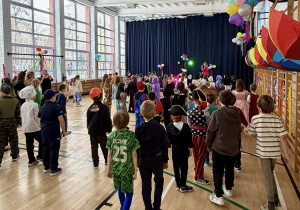 Uczniwoei w przebraniach stoją w udekorowanej sali gimnastycznej i słuchają powitania Pani animator.