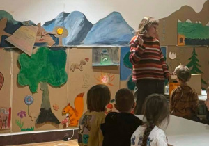 Pierwszoklasiści, siedząc przy stolikach, uważnie słuchają na temat historii zabawek.