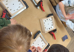 Uczniowie, siedząc przy stole, wykonują zegary z drewna.