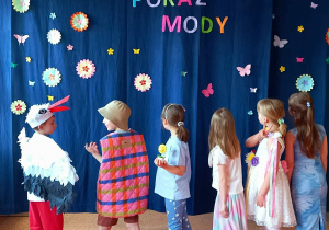 Uczniowie prezentują piękne i kolorowe stroje stojąc na tle napisu dekoracyjnego "Wiosenny Pokaz Mody".