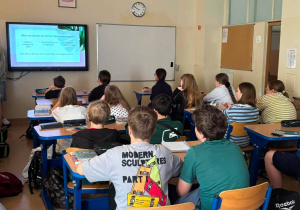 Uczniowie siedzą w ławkach i oglądają prezentację.