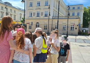 Dzieci stoją na Krakowskim Przedmieściu i słuchają przewodnika.