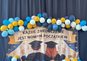 Dekoracja w sali gimnastycznej: baner z napisem "Każde zakończenie jest nowym początkiem" i kolorowe balony wiszą na zasłonach.