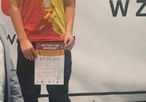 Na zdjęciu widać ucznia naszej szkoły, który stoi na podium i trzyma w rękach dyplom. Chłopiec ma na sobie czerwono-żółtą koszulkę, ciemne spodnie oraz medal zawieszony na szyi. Za nim znajduje się czerwono-białe tło z dużą koroną.
